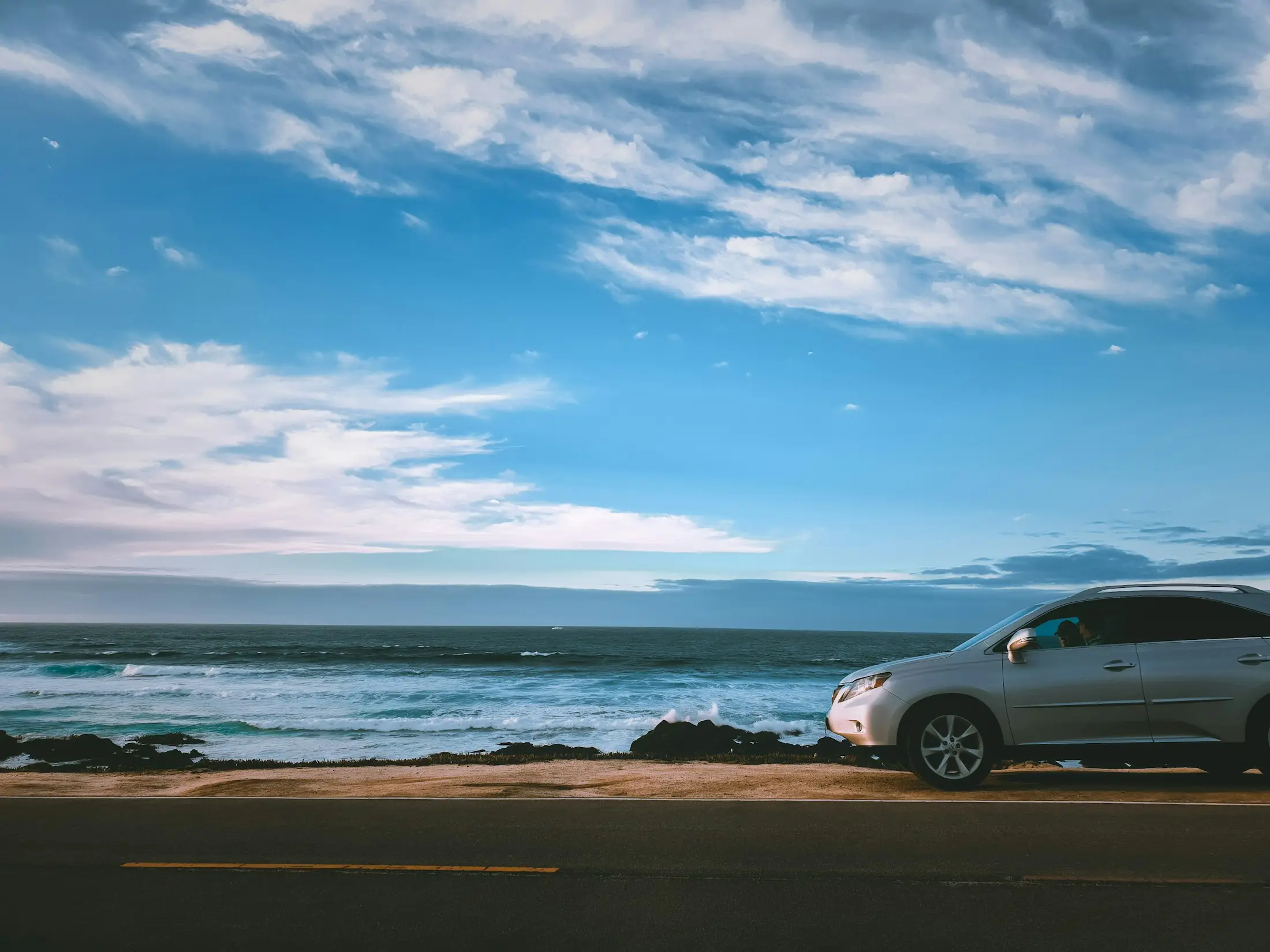 Silver SUV parked beside a coastal road with ocean waves and a blue sky behind it