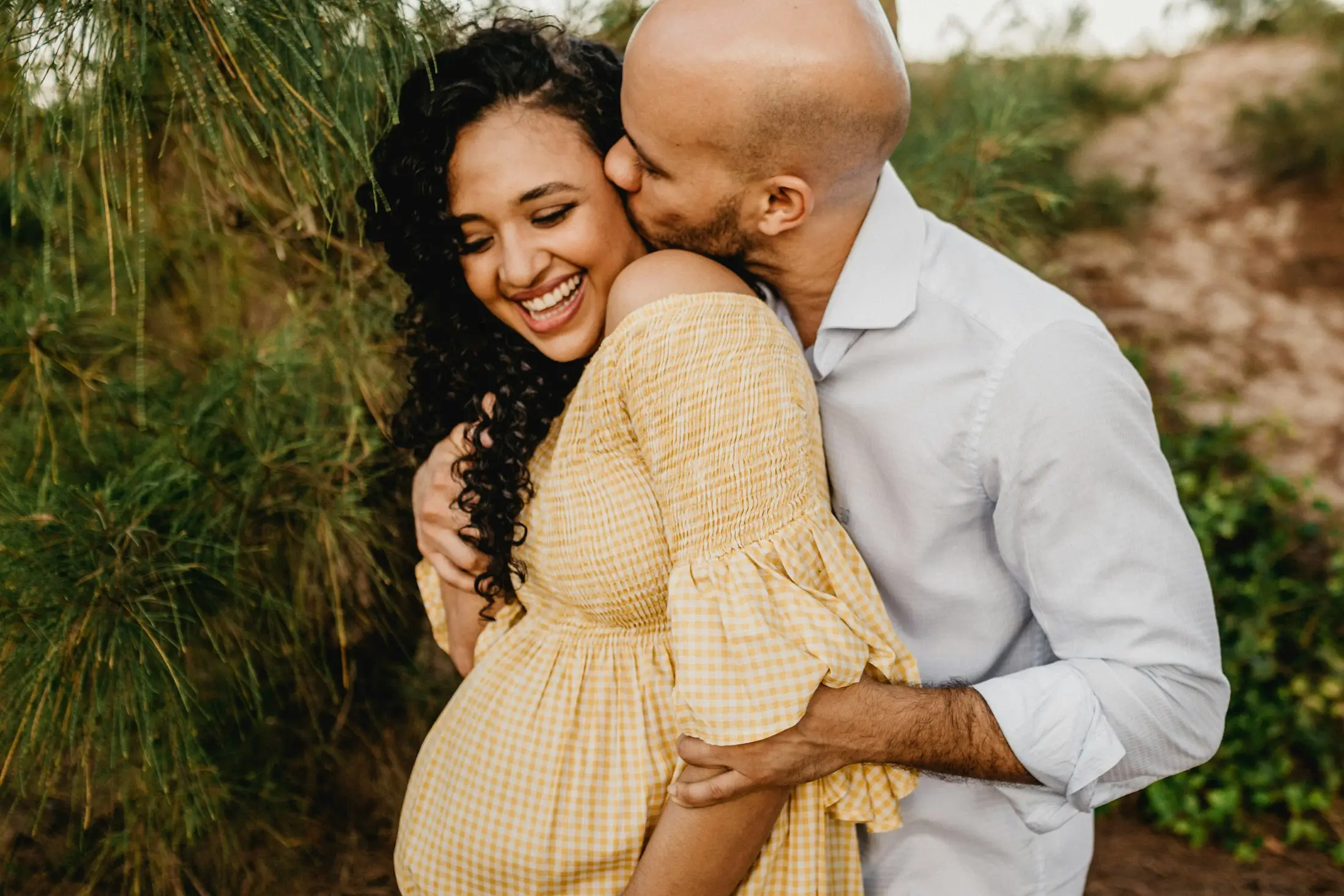 Smiling expectant couple embracing outdoors, the man kissing his partner’s cheek among green foliage