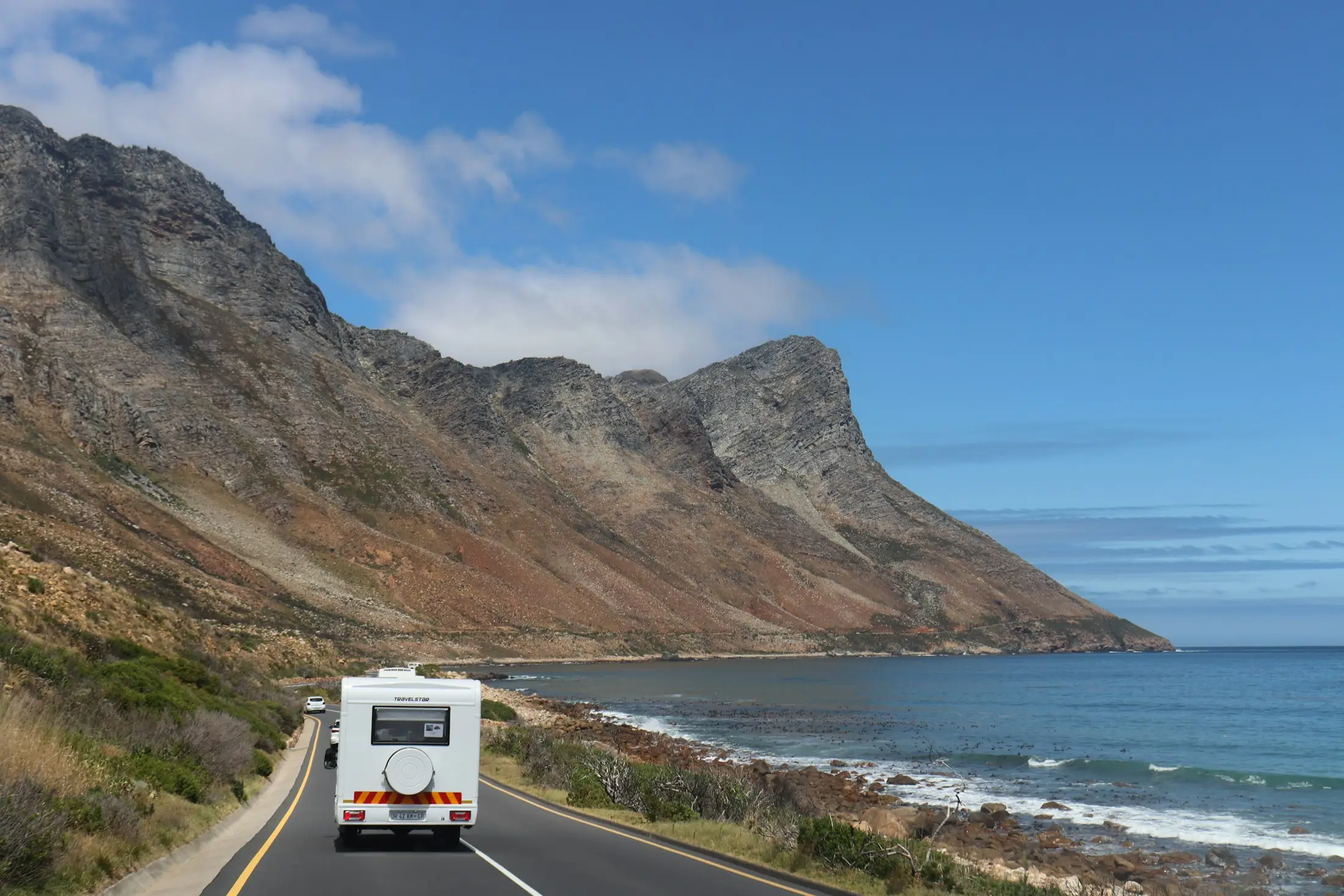 Motorhome driving a coastal road beneath dramatic mountains