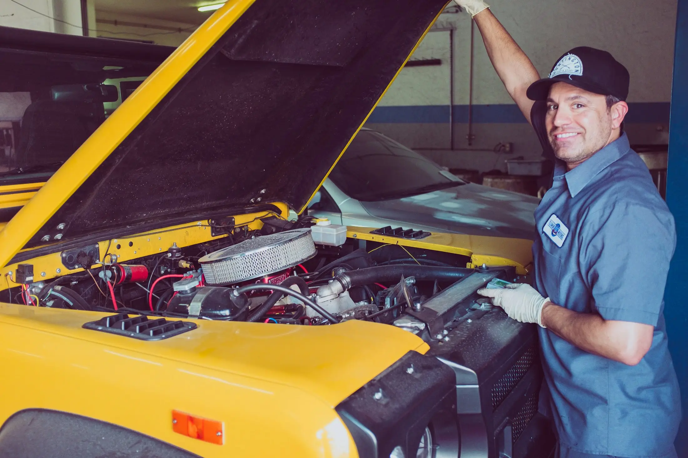 Smiling mechanic in a workshop holding up the bonnet of a yellow car