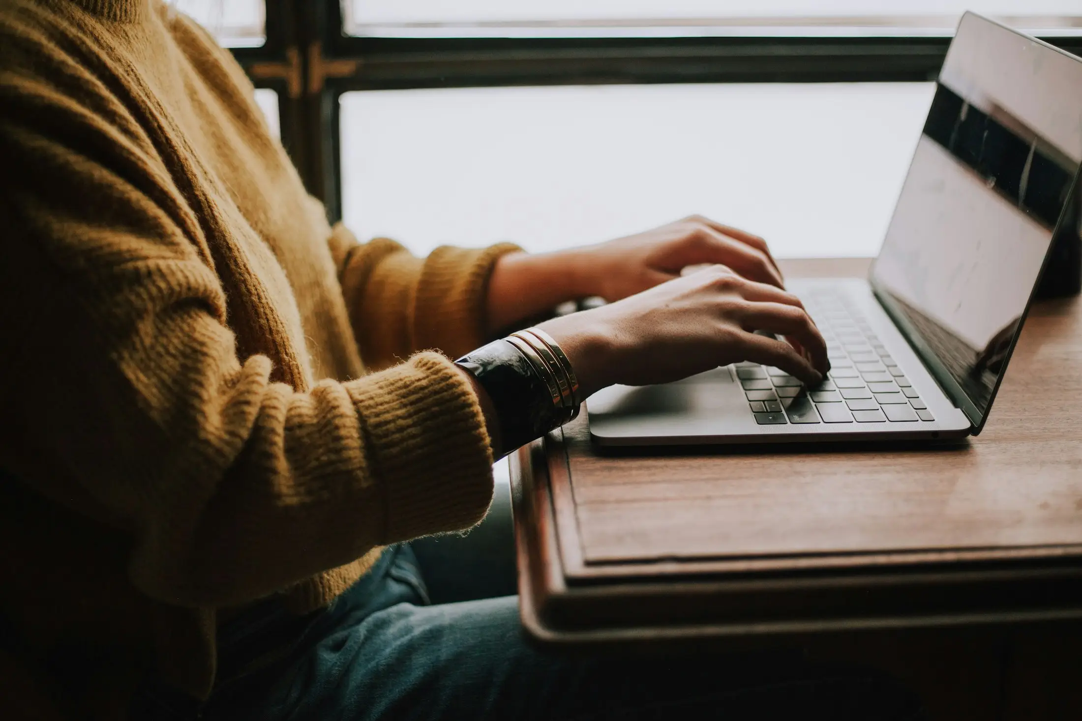 Person in a mustard jumper typing on a laptop at a wooden desk while gathering documents online